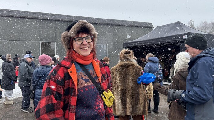 Une femme souriante au premier plan porte une tuque en fourrure et une veste à carreaux rouges. Elle se trouve à l'extérieur avec d'autres personnes devant un bâtiment gris et une tente noire, sous une légère chute de neige.