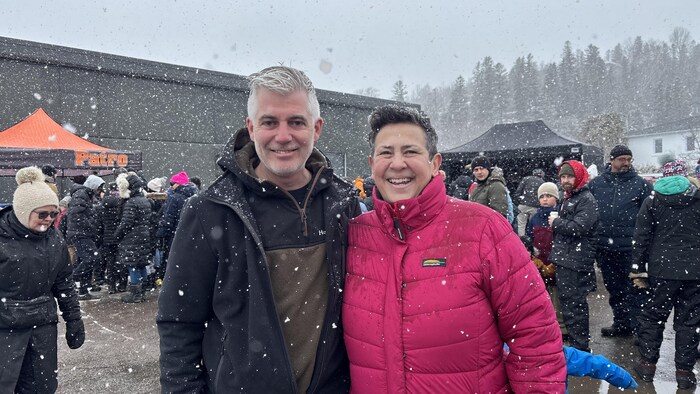 Un homme en manteau noir et une femme en manteau rose fuchsia sourient au premier plan. Ils participent à un rassemblement extérieur sous une neige abondante, entourés d'une foule, près d'un bâtiment gris et de tentes.