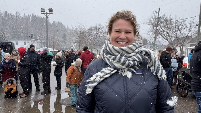 Une femme souriante au premier plan porte un manteau matelassé bleu foncé et une écharpe rayée. Derrière elle, une foule est rassemblée à l'extérieur sous la neige, avec des arbres et des maisons en arrière-plan.