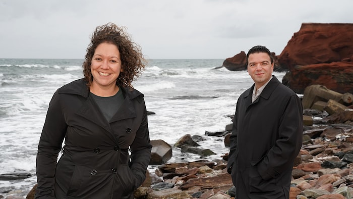 Une femme et un homme sourient à la caméra devant une mer agitée et des falaises aux Îles-de-la-Madeleine.
