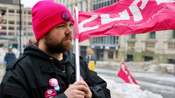 Un homme regarde vers la droite d'un air déterminé. Il tient à deux mains le manche d'un drapeau rose avec les lettres CUPE.