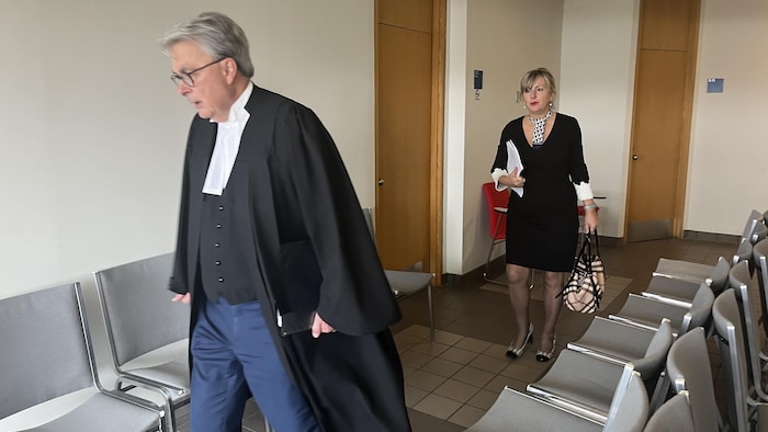 Un homme en habit d'avocat marche dans un couloir suivi d'une femme tenant des documents.