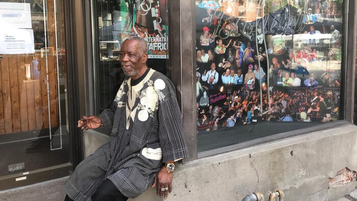 Un homme fume la cigarette alors qu'il est assis sur le rebord de la fenêtre d'un local commercial. Dans la vitrine sont présentées des photos de musiciens africains.