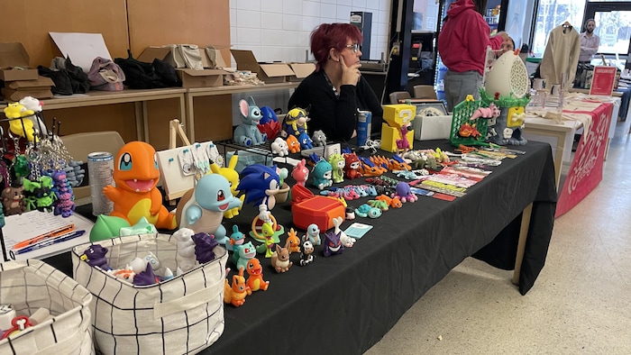 Une exposante aux cheveux rouges se tient derrière une table noire couverte de figurines colorées et de porte-clés. L'événement se déroule dans une salle publique avec d'autres kiosques et des visiteurs en arrière-plan.