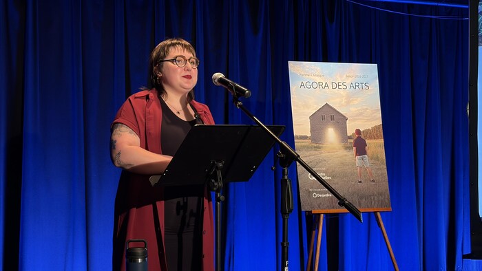 Becky Vander Haeghe parle au micro lors du lancement de programmation de l'Agora des Arts, à côté de l'affiche où l'on voit un jeune garçon, de dos, regardant une maison abandonnée.