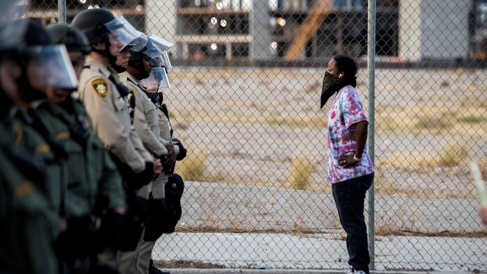 Une femme noire masquée se tient debout devant une rangée de policiers armés et casqués.