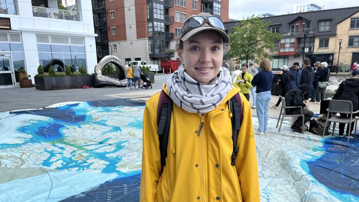 Une femme vêtue d'un imperméable jaune et coiffée d'une casquette sur laquelle des verres fumés ont été posés se tient droite sur une carte géante du Canada posée sur le sol, en public.
