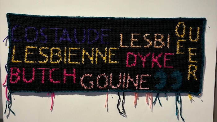 Une tapisserie où sont écrits les mots "costaude", "lesbienne", "lesbi", "queer", "butch", "dyke", "gouine".