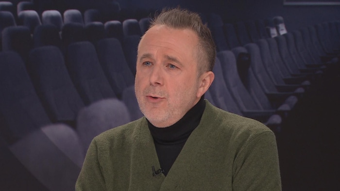 Laurent Dubois donne une entrevue sur un plateau de télévision devant une image de sièges dans une salle.