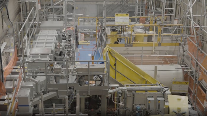 Un petit couloir passe au milieu de machinerie complexe. Le symbole de la radioactivité est présent sur deux pancartes.