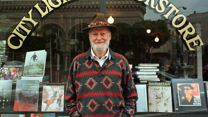 Lawrence Ferlinghetti, qui porte un chapeau brun et une barbe blanche, est debout devant la vitrine de sa librairie. 