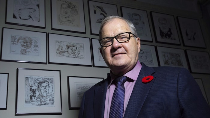 Lawrence MacAulay debout devant un mur auquel une douzaine de cadres sont accrochés, représentant des caricatures qui ont été faites de lui par des illustrateurs.