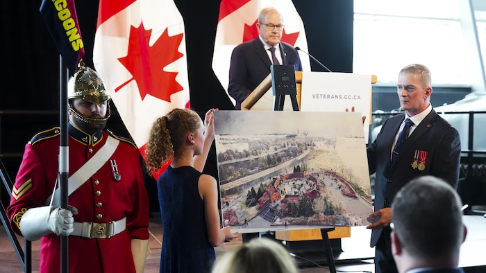 L'ancien ministre des Anciens Combattants du Canada, Lawrence MacAulay, regarde le carporal Nick Hebert et sa fille Kassandra Hebert qui dévoile la maquette du design gagnant.