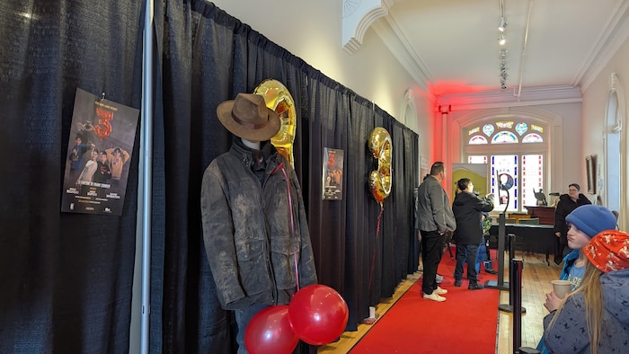 Dans un théâtre, une affiche de film et un mannequin sur lequel on a posé un manteau noir et un vieux chapeau brun sont posés sur un tapis rouge, le long d'un rideau noir.