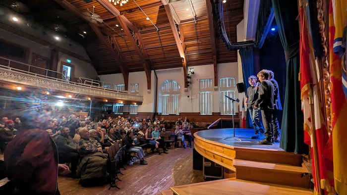 Trois enfants sur une scène, l'un parlant dans un micro, devant un théâtre rempli de spectateurs.