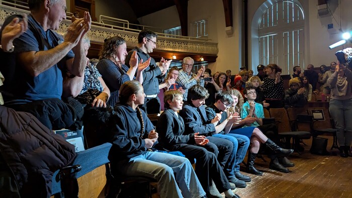 Dans un grand théâtre, trois enfants sont assis au premier rang. Les personnes à l'arrière se sont levées et ils applaudissent.