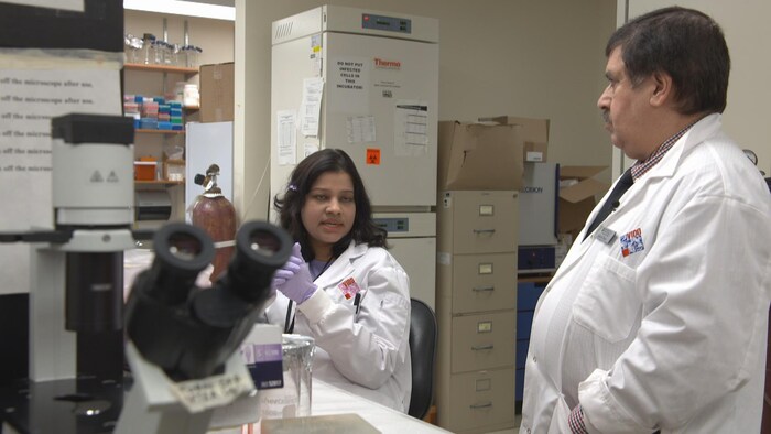 Suresh Tikoo, à droite, qui porte un sarrau de laboratoire, parle à une chercheuse, à gauche, qui porte aussi un sarrau. Il y a un microscope à l'avant-plan.