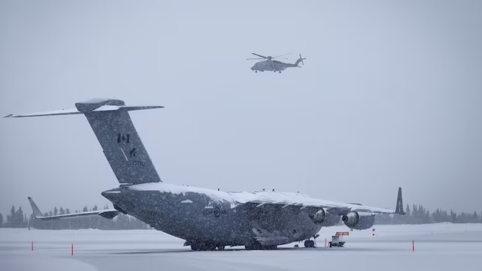 Un avion militaire enneigé sur une piste, avec un hélicoptère qui vole au-dessus.