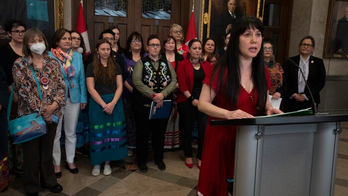 Une femme vêtue d'une robe rouge se tient lit un discours devant un podium. Derrière elle, des militants pour les droits des Autochtones et des drapeaux du Canada. 