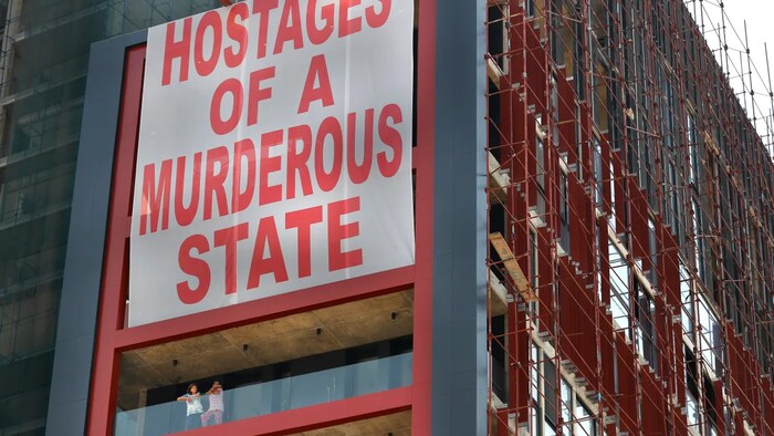 Citizens stand on their apartment balcony under a giant banner that hangs on a building damaged during last year's seaport explosion during a one year commemoration in Beirut. (Hussein Malla/The Associated Press)