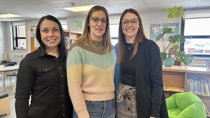 Trois femmes debout et souriantes posent côte à côte dans une bibliothèque scolaire. Elles sont placées devant des étagères de livres et une plante verte, dans un environnement lumineux comprenant des tables de travail au second plan.