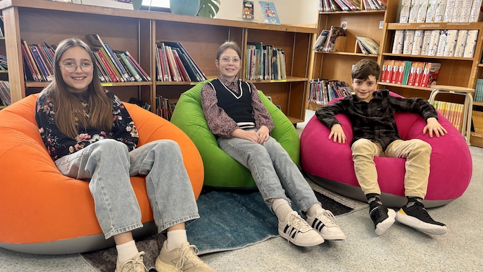 Trois enfants souriants sont assis côte à côte dans des poufs colorés (orange, vert, rose) devant des étagères remplies de livres. Ils regardent l'objectif dans ce qui semble être une bibliothèque scolaire ou un coin lecture.