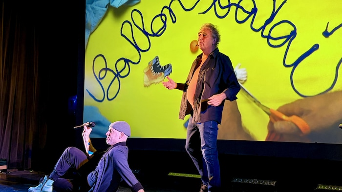 Dominiq Hamel est assis sur scène, pendant que Pierre Labrèche performe un conte devant un film d'animation.