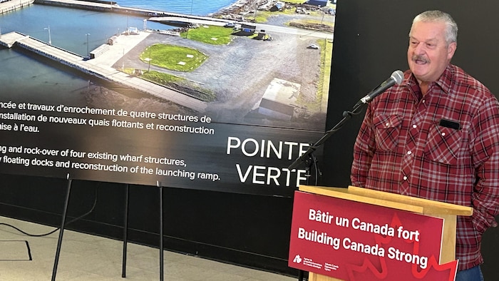 Léger Arseneau parle à un micro devant une pancarte avec le port de Pointe-verte.
