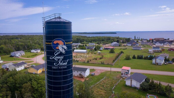 Un réservoir d'eau peint en bleu avec un logo et le nom de Lennox Island surplombe des habitations et des terres l'été.