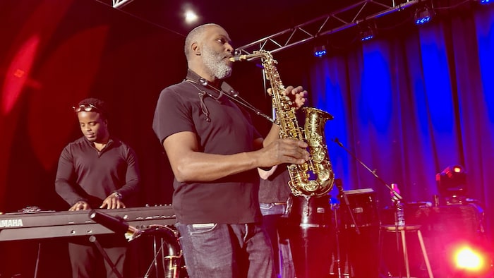 Le profil gauche d'un saxophoniste qui joue de son instrument sur scène, dans une lumière rouge et bleue, avec un claviériste jouant derrière.