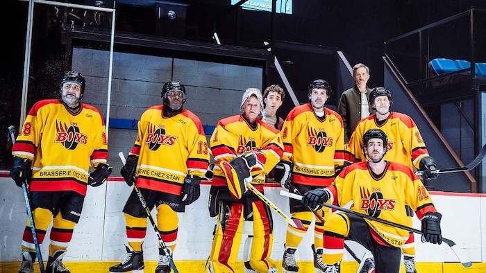 Six joueurs de hockey de l'équipe des Boys sont sur la glace et deux hommes sont debout derrière eux.