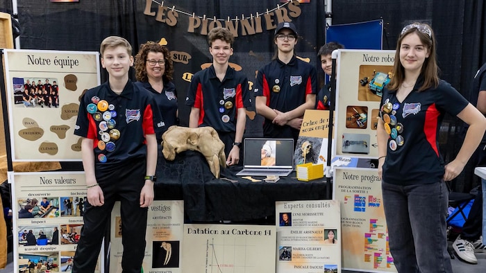 Autour d'un kiosque bien rempli sur les artéfacts, cinq adolescents sourient à l'objecitf, accompagnés de leur enseignante.