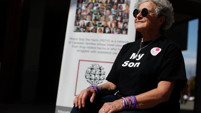 Une dame âgée avec des verres fumés et un t-shirt noir sur lequel il est écrit en anglais : mon fils, est assise devant une affiche avec plusieurs photographies de visages.