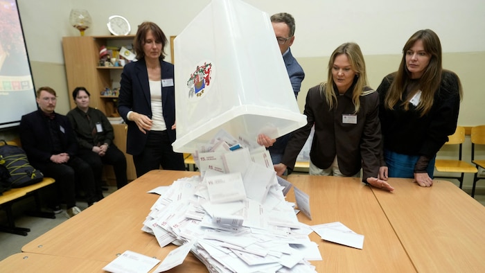 Entouré par trois responsables électorales, un homme renverse sur une table les bulletins de vote d'une boîte de scrutin, observé par deux autres personnes assises derrière.