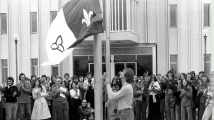 Vieille photo en noir et blanc d'un lever de drapeau devant une foule qui applaudit.