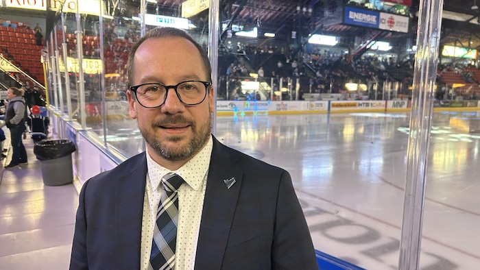 Un homme en complet sourit à la caméra devant une patinoire. Les gradins en arrière-plan sont à moitiés pleins.