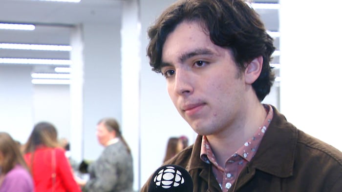 Un jeune homme debout dans une salle de conférence avec un micro de Radio-Canada devant lui.