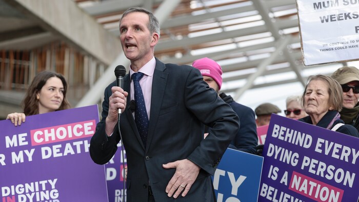 Liam McArthur debout, micro à la main, devant des manifestants tenant des pancartes exprimant leur appui à la pratique de l'aide médicale à mourir.