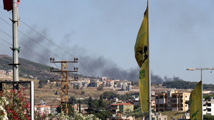Fumée noire s'élevant au-dessus d'une colline d'habitations, derrière un pylône électrique et un drapeau jaune.