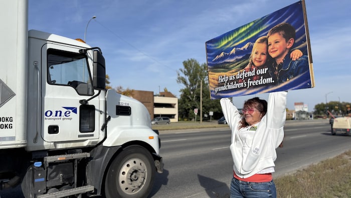 Une femme tient une pancarte incitant les automobilistes à klaxonner. Il y est écrit : « Aidez-nous à défendre la liberté de nos petits-enfants. »