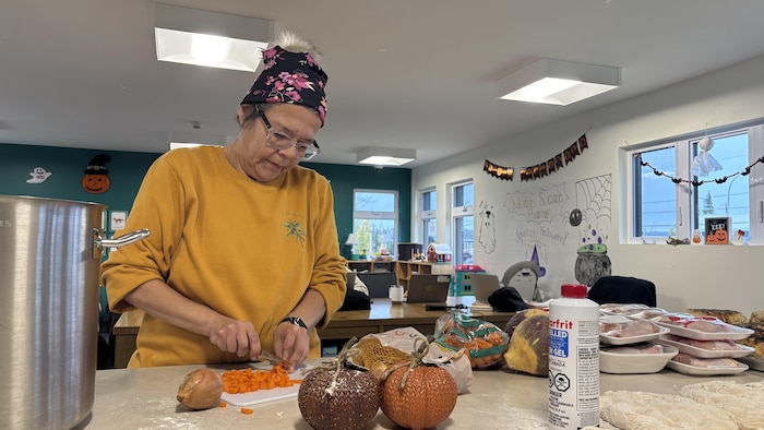 Liette St-Onge, portant un bonnet et des lunettes, est concentrée à couper des carottes en petits dés sur une planche à découper. Sur le comptoir devant elle se trouvent une grande marmite métallique, des oignons, un navet, plusieurs barquettes de viande ainsi que des décorations thématiques.