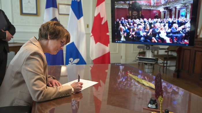 La lieutenante-gouverneure signant une nomination devant une télévision allumée.