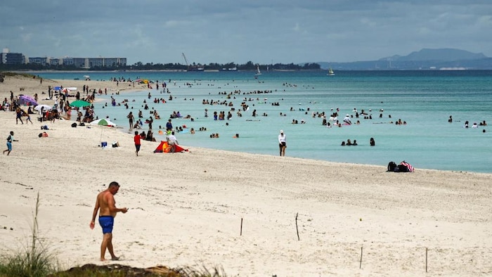 Des touristes profitent de leurs vacances sur une plage de sable blanc devant une mer bleu claire. 