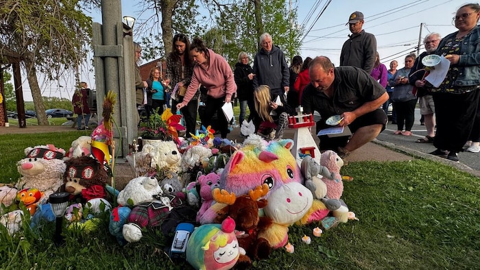 Plusieurs personnes agenouillées dans la pelouse, où se trouve un mémorial composé de fleurs et de peluches pour enfants.