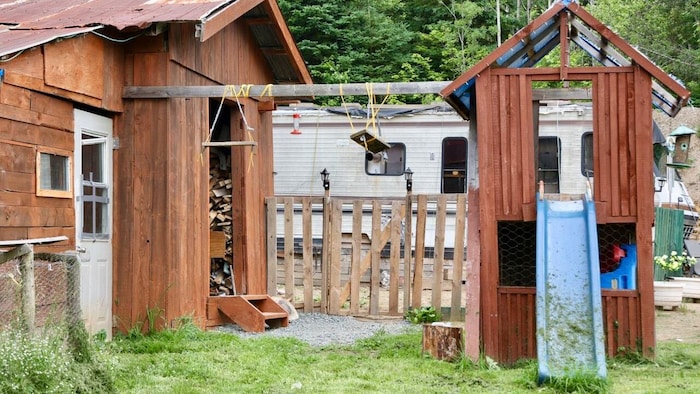 Sur une propriété rurale, deux balançoires sont enroulées autour d'une poutre horizontale reliant un cabanon à une structure de jeu pour enfants d'où descend une glissoire bleue.