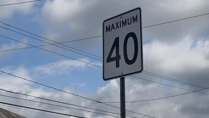 Un panneau de signalisation routière rectangulaire blanc, bordé de noir, affiche l'inscription « MAXIMUM 40 » en lettres et chiffres noirs. Il est fixé sur un poteau métallique face à un ciel nuageux traversé par plusieurs fils électriques.