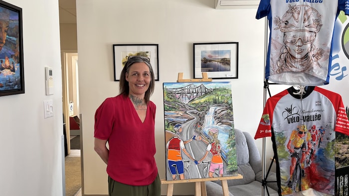 Linda Isabelle pose à côté de sa peinture qui met en scène des cyclistes sur une route près d'une chute d'eau.