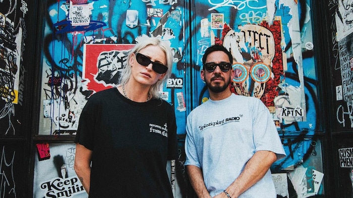 Emily Armstrong et Mike Shinoda posent avec des verres fumés devant une porte couverte de graffitis. 
