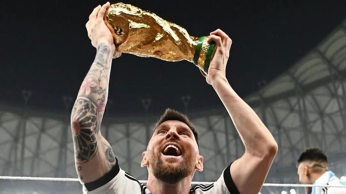 Un homme tient un trophée dans les mains qu'il porte au-dessus de sa tête, tout sourire, sur un terrain de soccer. 