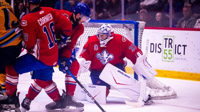 Un gardien se tient devant les buts alors que trois joueurs se trouvent devant lui.